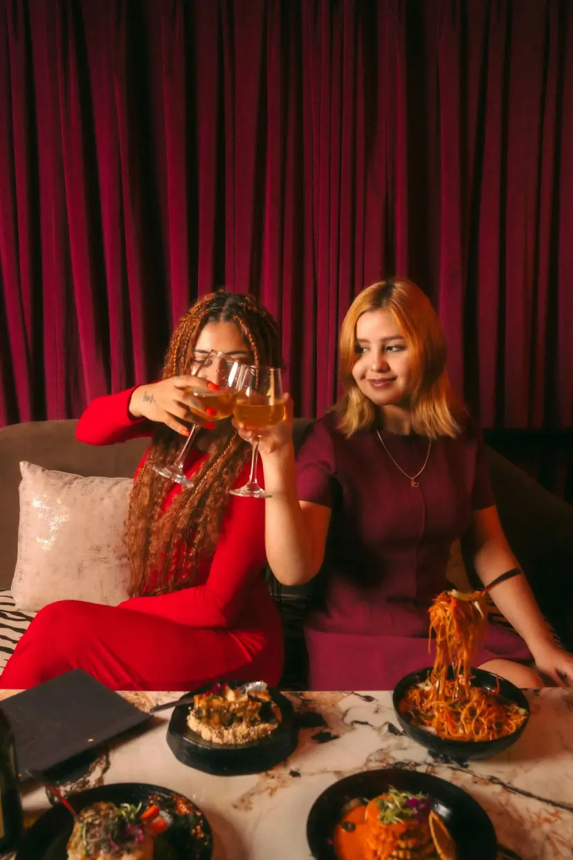 Two friends raise a toast over pasta in front of a red curtain.