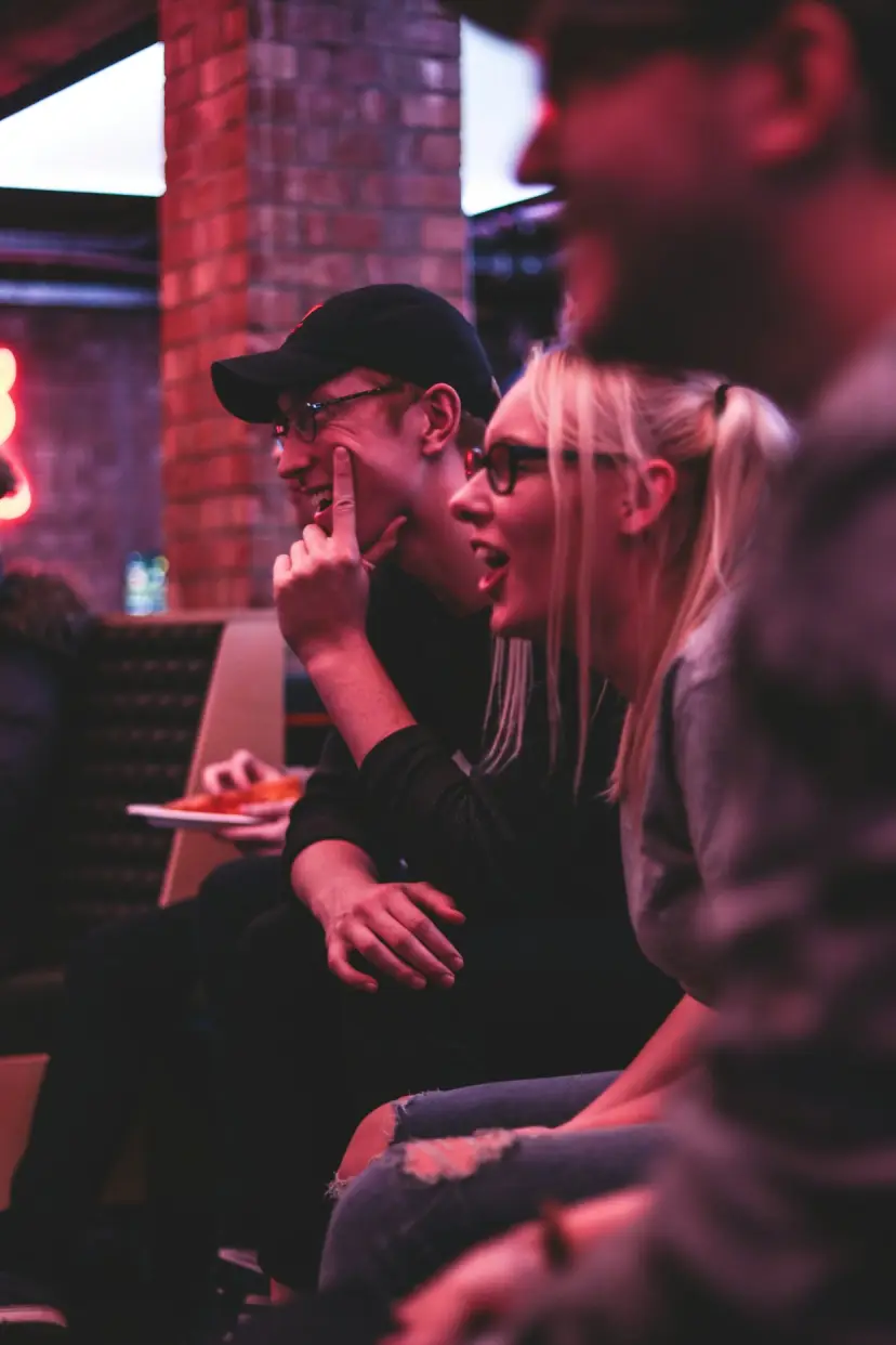 Friends laugh in a bar with a brick wall and neon light.